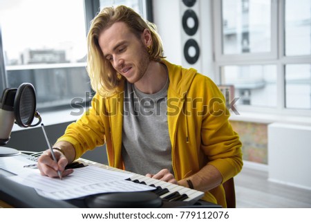 Portrait of cheerful young man composing melody with inspiration. He is writing notes on paper near electric piano and smiling