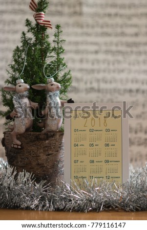 Calendar of 2018 year, a green plant and New Year's winter toys are on a wooden table. The calendar has words "Happy New Year 2018". Vertical postcard. 