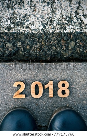 high angle view of four wooden numbers forming the number 2017 as the new year, and the feet of a man wearing blue shoes stepping on the asphalt