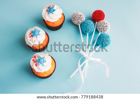 Sweet vanilla cupcakes with caramel filling and blue and white cheese cream, decorated with snowflakes and red, blue and white cake pops on colorful background.