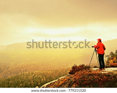 Professional photographer on location takes photos with mirror camera on peak of rock. Dreamy foggy landscape, spring orange pink misty sunrise in a beautiful valley below.