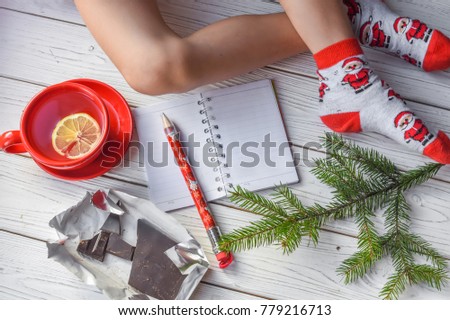 A festive Christmas photo with feminine feet in bright red socks, a fir twig, a note book and a pencil on a white wooden floor 
