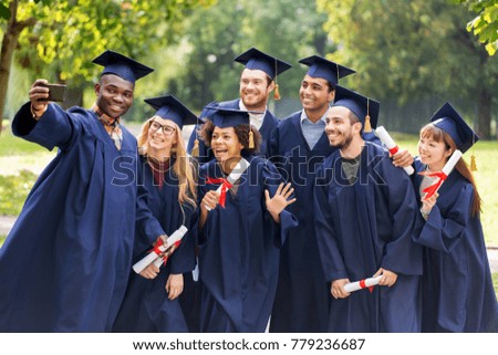 education, graduation, technology and people concept - group of happy international students in mortar boards and bachelor gowns with diplomas taking selfie by smartphone outdoors