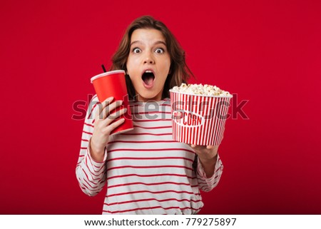 Portrait of an astonished woman holding popcorn and plastic cup isolated over pink background