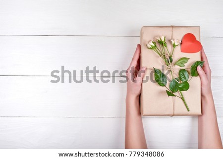 Woman holding present with fresh rose and heart on wooden background, free space. Close up of gift box in girl's hand, copy space. Holiday background. Flat lay, top view. Gift wrapping background