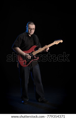A man in black clothes with an guitar in his hands is playing and posing on a black background with a blue light