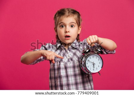 Picture of shocked little girl child standing isolated over pink background. Looking camera holding clock alarm.