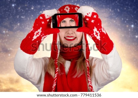 A young woman in a knitted hat scarf and mittens takes pictures of herself on the phone in a winter forest

