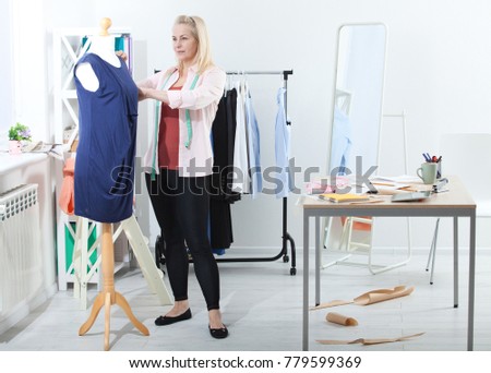 The concentrated work of tailor over details of dress. Beautiful European woman completes dress on mannequin, standing near  table full-length in workshop with clothes. Interier design studio.