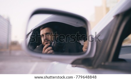 Reflection in side mirror of young private detective man sitting inside car and photographing with dslr camera