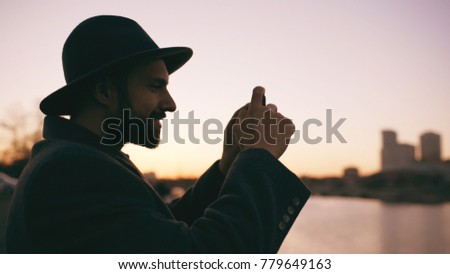Silhouette of traveler man in hat taking panoramic photo of the city skyline on his smartphone camera at sunset