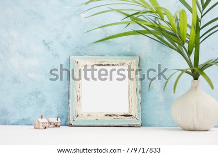 Square blue Photo frame mock up with green tropical plants in vaseand small wooden houses on shelf. Scandinavian style. Text space