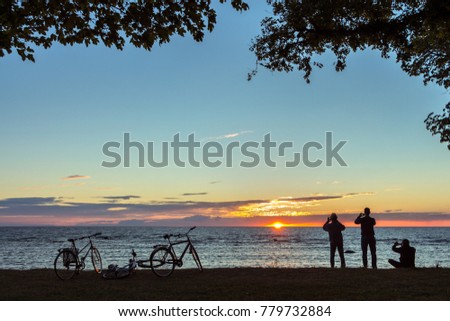 Sunset over Baltic sea in Visby on island of Gotland in Sweden. Beautiful colours, gradients and nature calmness.  Silhouettes of three men taking photos of sun and their bikes lying on grass. Summer.