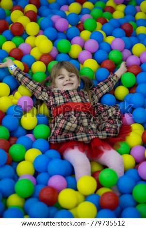 Happy little kid girl in red dress play in pool with colorful plastic balls. Funny child having fun indoors.