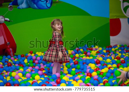 Happy little kid girl in red dress play in pool with colorful plastic balls. Funny child having fun indoors.