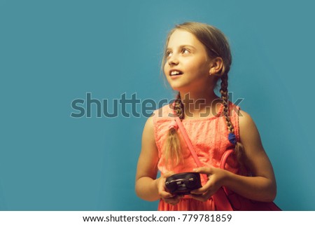Back to school and childhood concept. Kid with distracted face expression. Girl with black alarm clock and pink bag. Pupil with braids, isolated on blue background with copy space.
