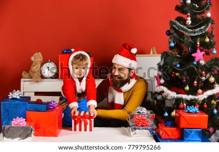 Winter holiday concept. Santa and little assistant among gift boxes near fir tree. Man with beard and smiling face plays with son on New Year eve. Christmas family opens presents on red background