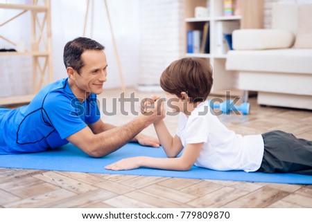 The man and the boy are engaged in arm-wrestling. This is father and son. They are at home.