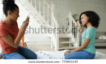 Cheerful mixed race woman photographing his girlfriend using smartphone camera sitting on bed in bedroom at home