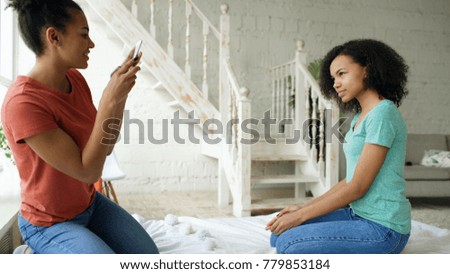 Cheerful mixed race woman photographing his girlfriend using smartphone camera sitting on bed in bedroom at home