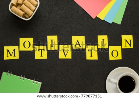 Motivation horizontal inscription. Checkered letters printed on yellow papers, blank notebook, coffee, cookies and note papers. Black concrete background. Space for your text or image. Top view.