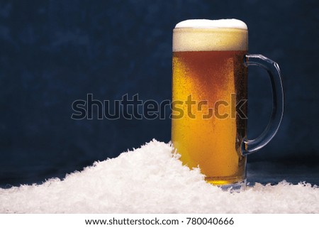 A glass mug of foaming light beer in the snow on a dark background. The theme of winter holidays.