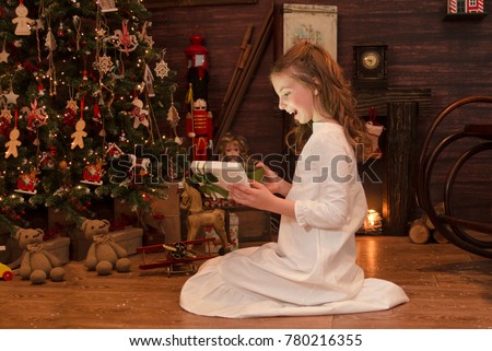 Excited little girl open gift box on Christmas morning. Smile and happy child look surprised at New Year present,siting by the Christmas tree. Kneeling on  floor in decorated vintage cottage.