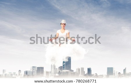 Woman in white clothing keeping eyes closed and looking concentrated while meditating on cloud above wooden floor with cityscape view on background.