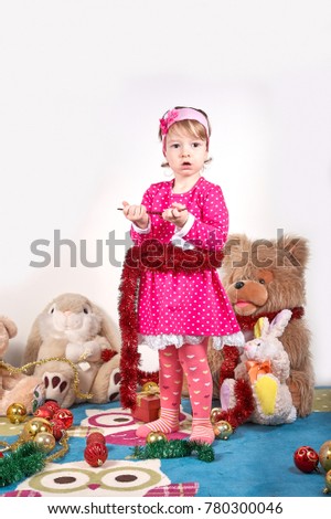 Adorable happy little girl kidin a red dress and a head band made of flowers is sitting on the carper and playing with Christmas toys