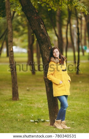 Beautiful happy caucasian young smiling brown-hair woman in yellow coat, jeans, boots in green forest. Fashion female model with fall golden leaves standing and walking in early autumn park outdoors
