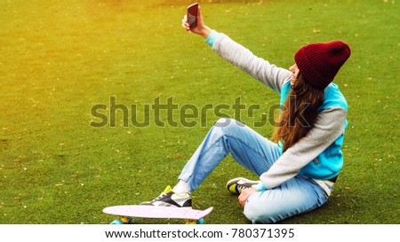 the girl is taking pictures of herself on the phone. sits on the grass. penny board. good weather