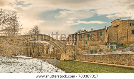 river placidly flows under an ancient donkey back bridge connecting medieval village to snowy countryside in Italy