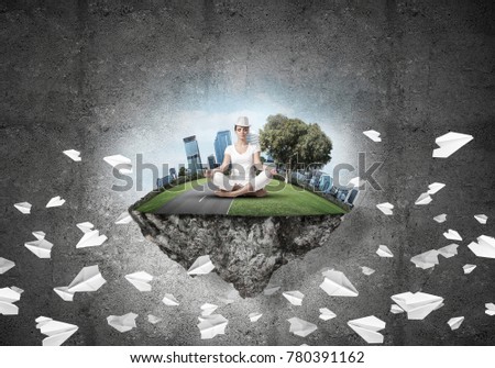 Woman in white clothing keeping eyes closed and looking concentrated while meditating on island in the air among flying paper planes with gray dark wall on background.