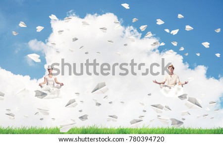 Young couple keeping eyes closed and looking concentrated while meditating on clouds among flying paper planes with bright and beautiful landscape on background.