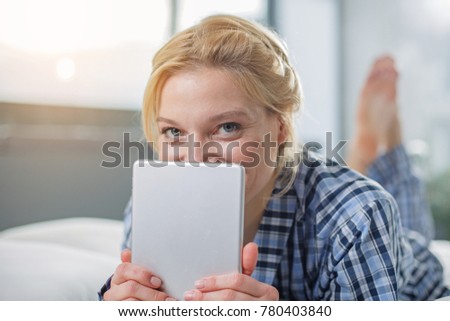 Portrait of happy girl resting on cozy beddings. She is holding tablet in front of her face