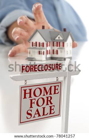 Foreclosure Real Estate Sign in Front of Woman's Hand Reaching for Model House on a White Surface.