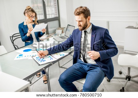 Portrait of a businessman dressed in suit working on documents with female assistant on the background at the white office interior