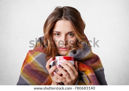 Winter cold sickness concept. Freezing beautiful woman wrapped in warm checkered plaid blanket, drinks hot beverage, tries to warm herself after spending time outside, home warm cozy atmosphere
