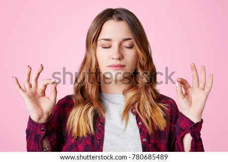 Beautiful female makes mudra sign, practises yoga indoor, meditates and tries to concentrate, enjoys peaceful atmosphere, isolated over pink studio background. Focused pretty female indoor