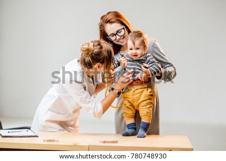 Mother with her baby boy at the young female pediatrician listening with a stethoscope standing at the white office interior