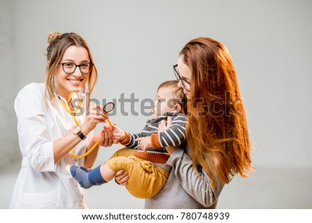 Mother with her baby boy at the young female pediatrician listening with a stethoscope standing at the white office interior