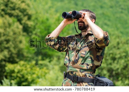 The smiling man in military uniform is watching with binoculars in nature on greenery background.