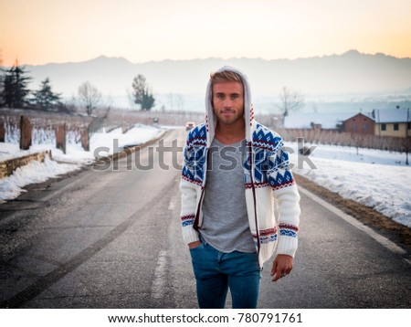 Handsome young man outdoor in winter fashion, wearing black coat and woolen scarf in snow environment on a country road