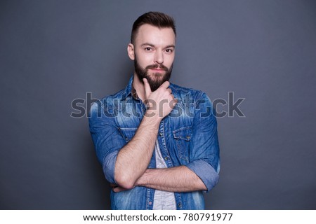 Young stylish smiling guy in a denim jacket looking at the camera and posing on a gray background.