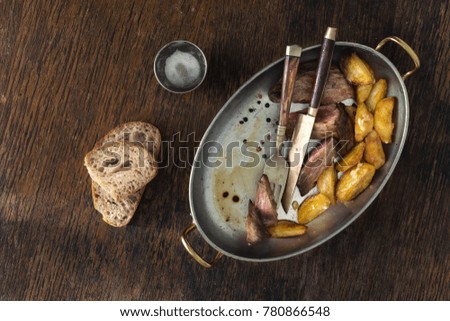Top view beef steak with fried potatoes in frying pan on dark background. Dinner table concept