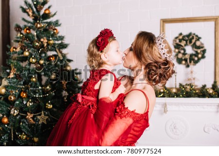 Young beautiful mother holding her infant daughter both wearing red dresses, kissing and hugging by the Christmas tree