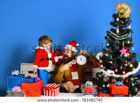 Santa and little assistant hold old clock showing midnight. Christmas time concept. Christmas family waits for New Year on blue background. Boy and man with beard and happy faces celebrate Christmas