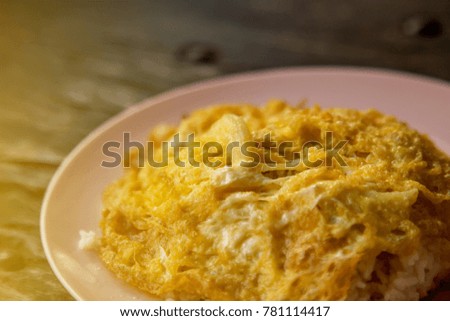 The fried omelette with white onion covering white jasmine rice in the pink plastic plate on the wooden table in the low light and flare from the picture corner.