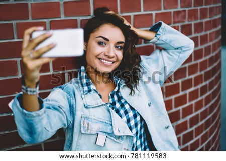 Attractive young woman with gorgeous curly hair posing and tacking selfie pictures via new photo application for uploading on own website standing outdoors against wall promotional background