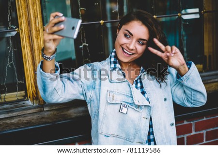Cheerful female blogger dressed in casual wear making funny selfie photos on telephone for uploading on internet webpage and sharing in social networks standing outdoors near architecture building